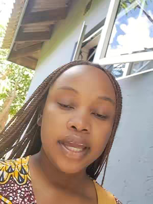 The cover image features a close-up of a woman with brown skin and braided hair. She is looking down with slightly parted lips as if in the middle of speaking. She is wearing a yellow and patterned top. The background includes a light-colored house and a partially visible window.