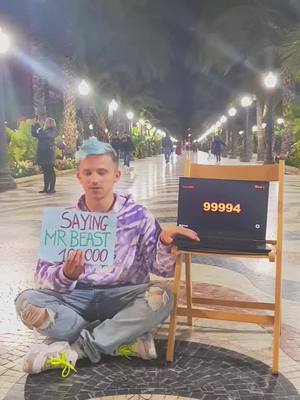 A young man with blue hair sits cross-legged on the ground holding a sign that says 'Saying MrBeast 10000'. In the background, a laptop with a lit display reads '99994' is propped on a wooden chair, and a walkway with lamp posts stretches into the distance. The composition is balanced, with the man centered and the ambient lighting suggesting an outdoor setting.