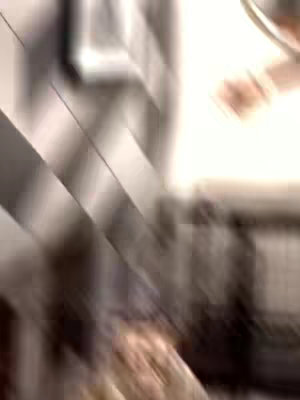 The image is a blurred, abstract view of a room or interior space. A diagonal gray and white striped pattern is prominent in the background suggesting a wall or similar structure. Bright, undefined light is visible on the right, hinting at a window or opening. The overall composition is out of focus suggesting movement.