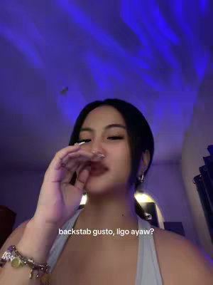 A young woman with dark hair is shown in a selfie with a casual background. She is holding her fingers up to her lips, in a gesture suggesting she is trying to contain or conceal something. The background is shaded in blue tones. The woman is wearing a light-colored top and jewelry. The text overlayed on the image is written in white font.