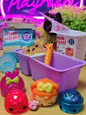 The image shows a colorful display of Num Noms toys arranged on a wooden surface. In the background, a neon sign that says "Playroom" in bright pink is visible. In the foreground, there are various Num Noms figures, including a pink bow-shaped one, an orange cupcake, and a blue ice-cream-shaped one, along with some of their accessories, all on a purple tub.  Various packages and clear blue plastic container are there too.  The overall tone is playful.