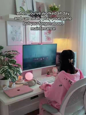 The image shows a woman with dark hair sitting at a computer desk. She is wearing a pink hoodie and sitting in a white chair. On the desk there's a white keyboard and a matching mouse. The background depicts a wall with framed pictures, a shelf with decorations, and a window with a curtain. She faces the computer monitor and has a well-lit, cozy setup.
