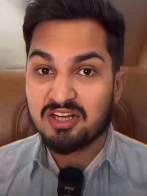 A medium shot of a man with a dark beard and slicked-back hair, speaking with an open mouth. He is wearing a light blue button-down shirt and has a microphone clipped to it. The background is blurred, and a hint of a brown surface is visible.