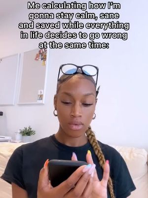 A woman with braided hair is holding a phone and looking at it. Text in white at the top reads "Me calculating how I'm gonna stay calm, sane and saved while everything in life decides to go wrong at the same time:". The background shows a partial view of a room with a cream-colored sofa and a plant.