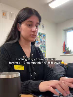 A young woman with long black hair is seated behind a desk, working on a laptop. Bold text overlays the image, providing a caption. The background is a generic office setting, with motivational lettering. The woman is wearing a black collared shirt.