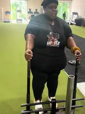A woman in fitness attire pushing a sled in a gym. The shot is taken from a low angle, emphasizing her physical effort. The background of the gym includes exercise equipment and windows.