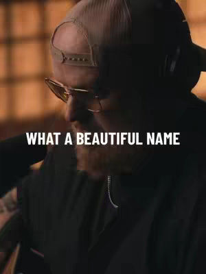 A man wearing a baseball cap and glasses with text "WHAT A BEAUTIFUL NAME" in white. The image has a dark, moody lighting style.