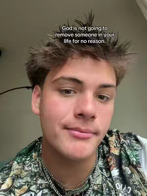 A young man with light skin and brown hair is centered in a selfie-style shot. He is wearing a camouflaged shirt, and a silver chain around his neck. The background is a blurred, neutral color. Text overlay at the top reads a faith-based message.
