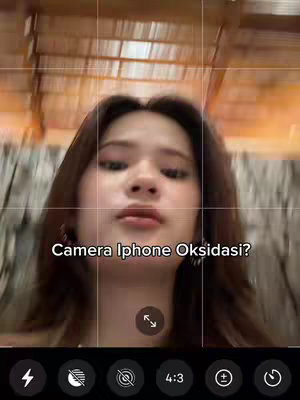 A selfie-style video shot with an iPhone camera interface visible, featuring a young woman at the center. The video is framed within a phone's recording interface, the woman is positioned in front of a blurred background. Text overlayed is  ‘Camera Iphone Oksidasi?‘ displayed in a clean, white font.