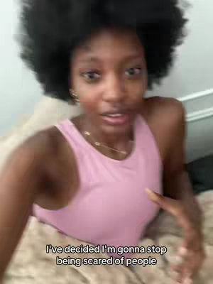 A selfie-style video shows a young Black woman in close-up, sitting in bed. She wears a pink tank top and necklace. Her dark, voluminous afro is prominent against a light-colored background. Text is overlaid at the bottom of the image.
