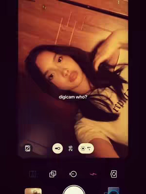 A woman with black hair takes a selfie. She is looking at the camera with a slight pout. A text overlay reads "digicam who?". The image has a warm tone and includes elements of a phone’s camera interface at the bottom.