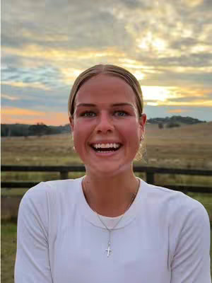 A young woman with blonde hair smiles widely at the camera against a beautiful sunset. She wears a white long-sleeved shirt and a necklace with a cross pendant. The background features a field and a fence.