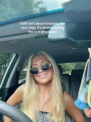 A selfie taken from inside a car. A young woman with long blonde hair and sunglasses smiles at the camera, sitting behind the steering wheel. The background is the car's interior and the outside environment. The image is well-lit, with a focus on the woman's face.