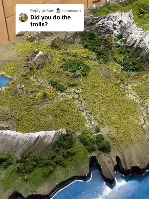 A high-angle shot of a detailed, miniature landscape model. The foreground is a detailed coastline transitioning into a landmass with varying elevations and textures. Lush green areas represent grassy terrains contrasting with gray and white mountain peaks. There's a small body of water to the left of the image. A text overlayed message in a white chat box on top left.
