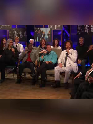 A stage is set with several men and women seated in chairs. The men are holding microphones. Behind them are various portraits and images. The lighting is a mix of blues and purples.