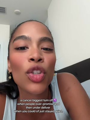 A selfie-style video shot of a woman speaking directly to the camera. She has brown hair, is wearing a light-colored tank top, and has makeup on. Text overlays are present near the bottom of the screen.