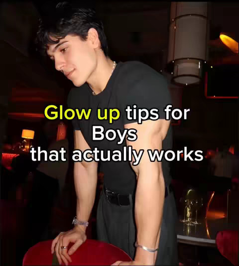 A young man in a black t-shirt is the focal point, set against a blurred indoor background with dim warm lighting. The overlay text suggests beauty tips for boys. The man is leaning on a red chair. He has dark hair and is looking down.