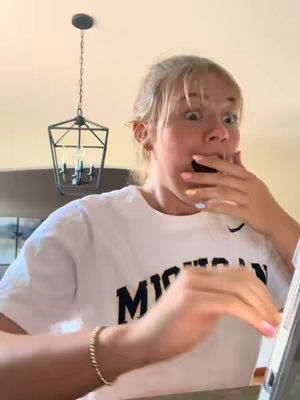 A young woman with blonde hair is captured from the chest up, reacting with surprise as she covers her mouth with her right hand. Her eyes are wide and her mouth is open in shock. She's wearing a white t-shirt that has a large black printed text 'MICHIGAN'. A gold bracelet is on her left wrist. The surrounding environment is simple with a light-colored ceiling which has a pendant light fixture hanging from it. There is background lighting along the top.