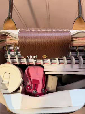 An overhead shot inside a light-colored, open tote bag. The bag is filled with organized study supplies: notebooks with white spiral bindings, a pink miniature backpack, a white cylindrical water bottle, and other items. In the foreground is a brown leather flap, embossed with a studded design, which covers part of the contents. The text 'studypee' in a dark font is placed centrally on the leather portion.