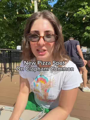 A woman wearing sunglasses is in focus, the text overlayed is about new pizza spot. Behind the woman the background is outdoors with a lot of green trees.