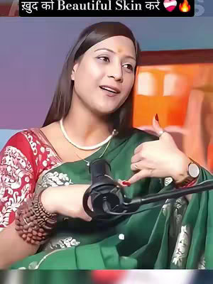 A woman is seated, speaking while looking slightly off-camera. She is wearing a green saree with a red floral patterned blouse. A pearl necklace and a wristwatch are visible. A microphone is positioned in front of her. Text in Hindi is overlaid across the top of the image. The background is softly lit, with hints of other objects and colors.