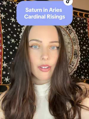 A woman with long, dark hair and light eyes is centered in the frame, with a light blue and white text box hovering above her head. The background is a tapestry with a celestial design.