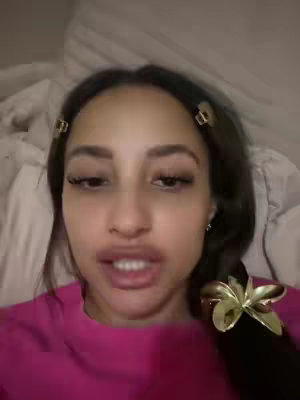 A close-up shot features a woman's face with a pink top and a white bedsheet in the background. She has her hair styled with gold hair clips and a clip on the ponytail. Her expression shows an open mouth with visible teeth.