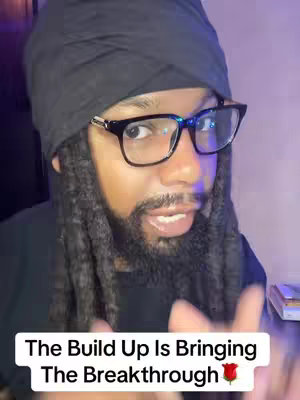 A man with locs and glasses looks at the camera with his index finger pointing up. Text at the bottom reads 'The Build Up Is Bringing The Breakthrough' with a rose emoji next to it.