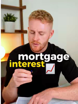 A man with fair skin and short, light brown hair is centered in the frame. He is wearing a black t-shirt and is set against a background of a white wall with wooden shelves and a small green plant. White and yellow text overlays read 'mortgage interest' with a red upward trending arrow icon next to it. The man appears to be speaking, gesturing with a pen in his hand over a document.