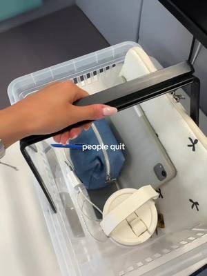 A hand holding a clear plastic bin with various items inside, including a blue pouch, a white cup, and a phone. The bin is on a white surface, with black trim visible.