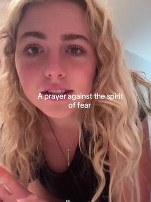 A close-up selfie of a young woman with long, wavy blonde hair. She is looking directly at the camera. The text overlay says, "A prayer against the spirit of fear". The background appears to be an interior space. She has a minimalist necklace on.