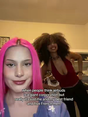 Two women are featured in a cover image, with one in the foreground and the other in the background. The woman in the foreground has bright pink hair and is centered on the left side of the image. She is wearing a blue top and facing the camera. The woman in the background has a large afro and is wearing a red top, appearing to be smiling. Text overlayed at the bottom of the image provides context. The backdrop appears to be a room corner with neutral-colored walls.