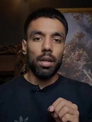 A man with a beard and dark hair is centered in the frame, looking at the camera with a slight expression. The background shows a scene of a painting with dark background.
