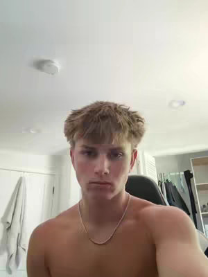 A shirtless young man with light brown hair and a silver chain necklace is centered in a vertical frame. He has a neutral expression. The background is a plain white ceiling with a light fixture and a glimpse of a wall with a door.
