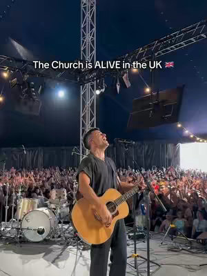 A live music performance taking place on a stage in front of a large crowd. The main focus is on a male musician playing an acoustic guitar and singing with great passion. The stage is well-lit with professional lighting rigs and screens at the back. The crowd is a large group of people, many with their hands up, captured in a wide shot from a slightly low angle. The overall impression is of energetic live music event.