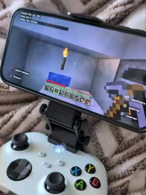 The image shows a smartphone displaying a Minecraft game, mounted on a black clip attached to a white Xbox controller. The phone is angled towards the viewer, showing a first-person view of a Minecraft scene within a gray-walled room. A torch is visible on the wall. The controller, with its buttons and joysticks, is at the bottom of the image. The background appears to be a bed. The lighting is warm, suggesting a cozy indoor environment.