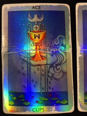 A close-up of a tarot card, specifically the Ace of Cups. The card has a holographic sheen, with a large golden chalice at the center. Water droplets cascade downwards. The background features a cloudy, dreamy effect and a body of water with lily pads at the bottom.