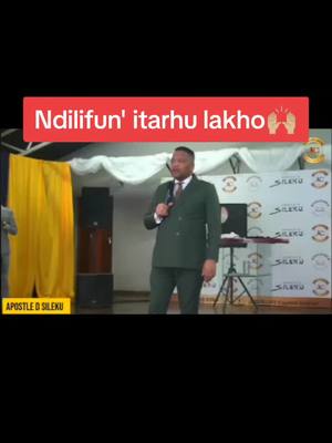 A man in a green suit is speaking into a microphone on a stage with a yellow curtain and various stage elements in the background. A red bar with white text is displayed above him. The background includes branding elements and speakers.
