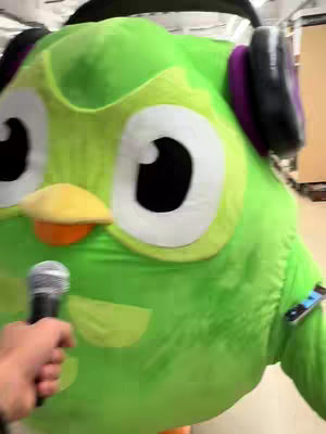 A person in frame is holding a microphone in front of a large green owl costume. The owl has large eyes, headphones, small yellow beak, and the body.