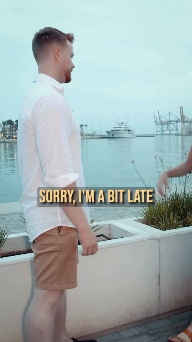 A man in a white shirt and khaki shorts stands on the edge of a waterfront, looking to the left. The text overlay, 'SORRY, I'M A BIT LATE,' is centered near the bottom. The background includes water, a distant harbor, and a cloudy sky.