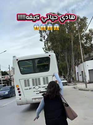 A woman in a dark vest and blue shirt is running away from a white bus, seemingly trying to catch it. The bus is angled slightly toward the viewer. The background includes buildings and trees under a cloudy sky.