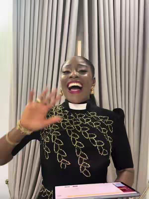 A woman with a radiant expression waves to the camera, set against a backdrop of vertical, beige curtains. She wears gold earrings and a black dress with ornate, gold embroidery across the bodice. A white collar suggests a clerical garment. Behind her is a laptop screen, potentially offering further context or information for the primary theme.