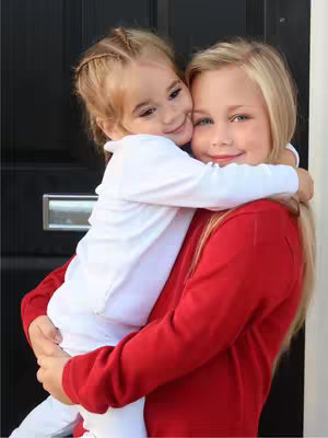 A young girl in a red long-sleeve shirt is holding a smaller girl in a white long-sleeve shirt and white pants. The smaller girl has blonde hair in braids. They are both smiling and hugging in front of a dark door.