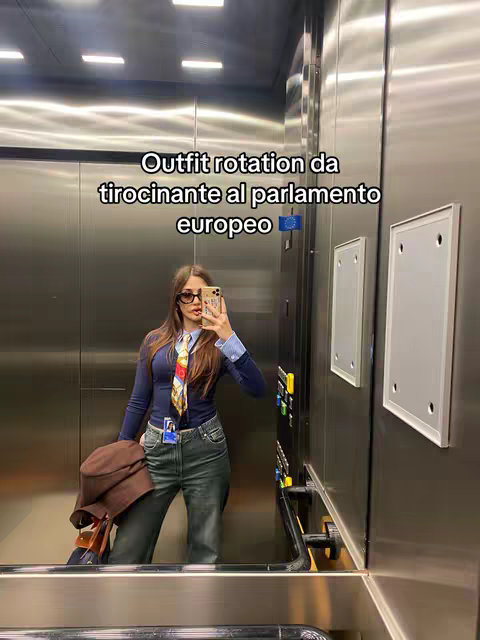 A person is shown in a well-lit elevator, using a phone to take a selfie. The person, who appears to be a woman, is wearing a navy blue top with a light-colored collared shirt underneath, jeans, a tie and glasses, and holding a brown hat, a sign of a formal attire. The background is the interior of an elevator, with metallic walls, showing a mirror reflection of the person holding their phone. Text overlay on the image indicates it's an outfit.
