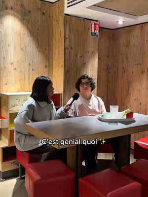 Two people are at a table, one holding a microphone and interviewing the other. The setting appears to be a restaurant or cafe with wooden paneling and red seating. A text overlay reads "C'est génial quoi" (It's awesome).