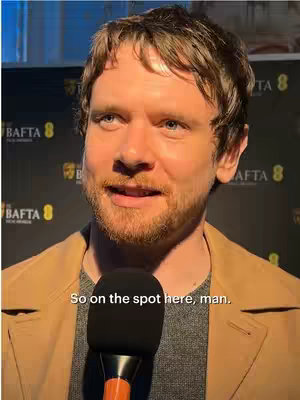 A man with light brown hair and light facial hair is shown close up, speaking into a black microphone. He wears a gray shirt under a tan jacket. The background has a repeating gold and black logo.