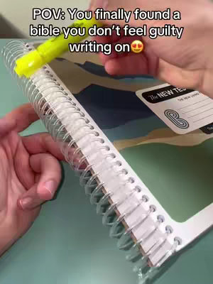 A person's hands are holding a yellow highlighter and using it to highlight text in a spiral-bound Bible. The bible is open showing the text and the spiral binding is clearly visible. The background shows a floral patterned surface.