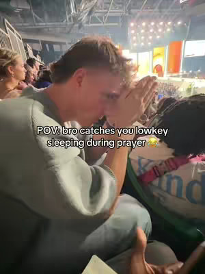 A selfie-style image of a young man in a crowd, seemingly at a religious event. He has short hair and is wearing a gray sweater. He has his hands together in a praying position, with his head bowed down. The background shows other people and stage lights, implying a large gathering. Overlayed text appears on the image.