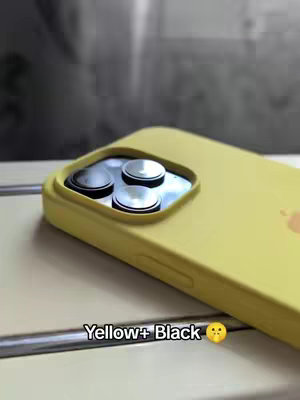 A close-up shot of a yellow iPhone angled slightly to the right, showcasing the camera lenses. The background is softly blurred and appears neutral. The text overlay at the bottom reads: \"Yellow+ Black \" with an emoji.