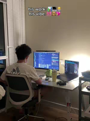 A man is sitting at a desk, facing away from the camera, working on a computer. The desk is cluttered with a laptop, monitor, drink, mouse, and other electronic devices. The image is dimly lit with a window off to the left.